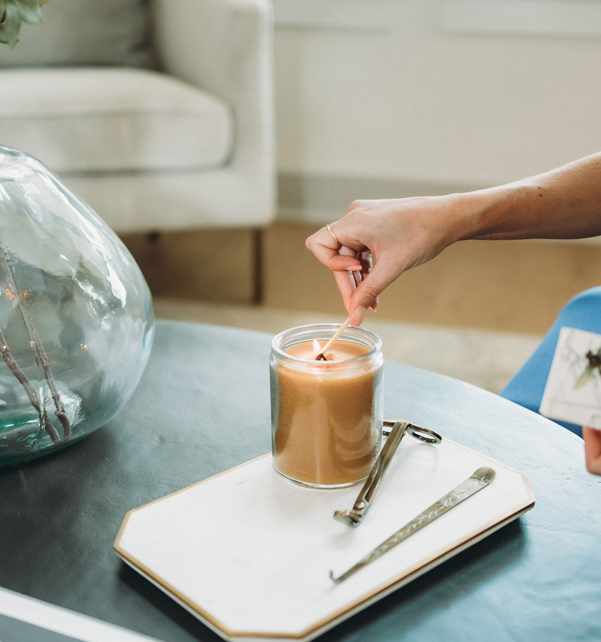 Person lighting a beeswax candle in a glass container on a table with candle accessories