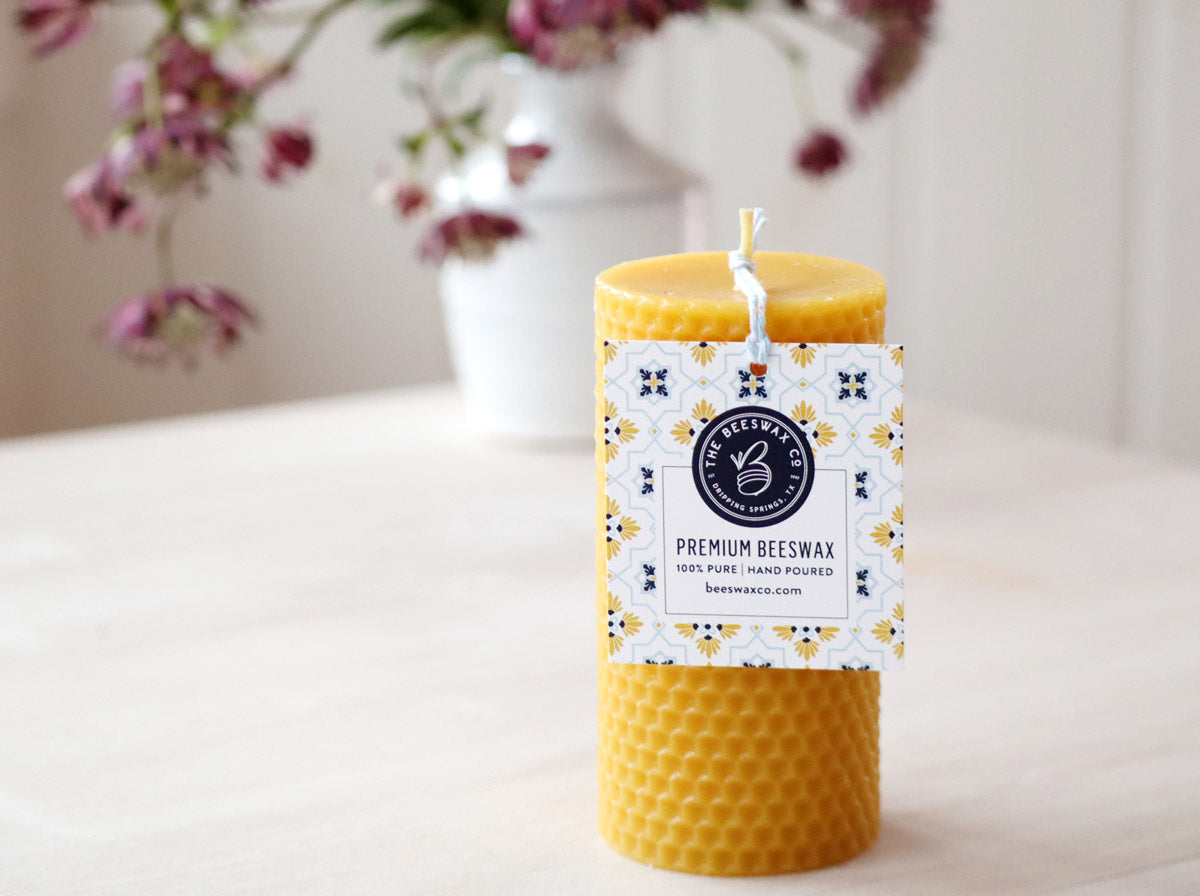 Beeswax candle with a label on a light surface with flowers in the background